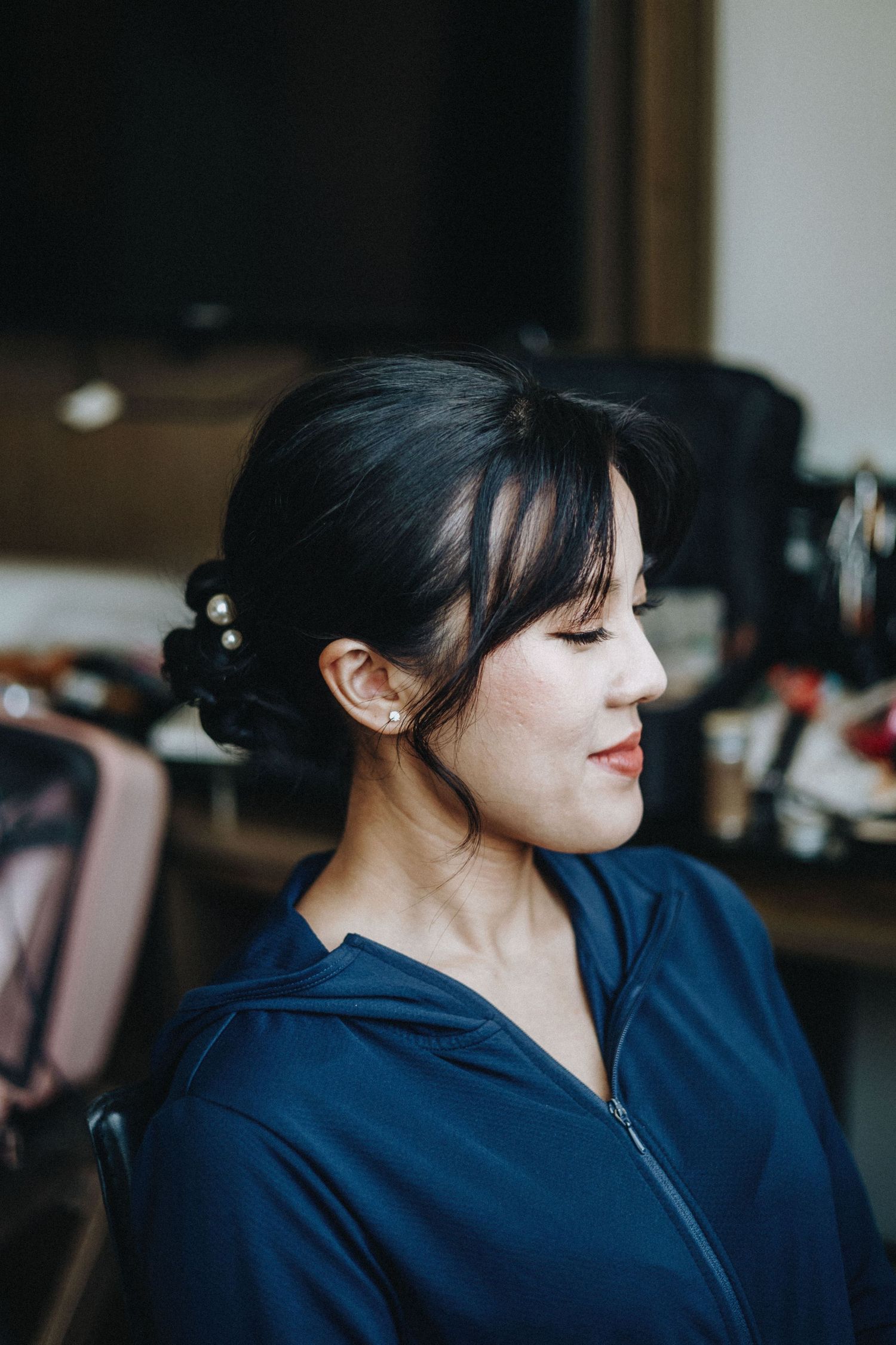 A woman with an elegant updo hairstyle and pearl hair accessories wearing a navy zip-up jacket, seen in profile view.