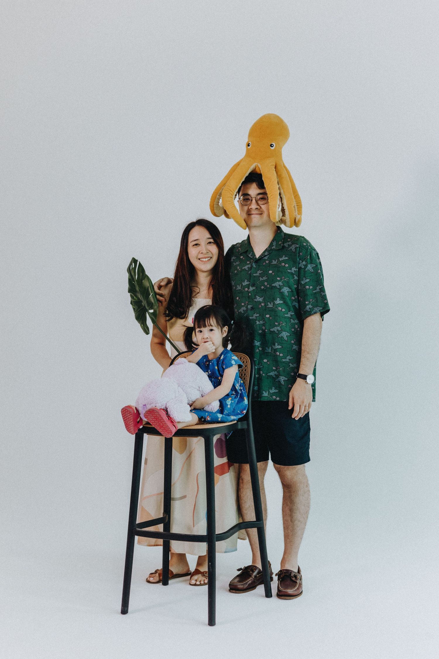 Family wearing matching green tones poses in studio with playful stuffed octopus and monstera leaf props.