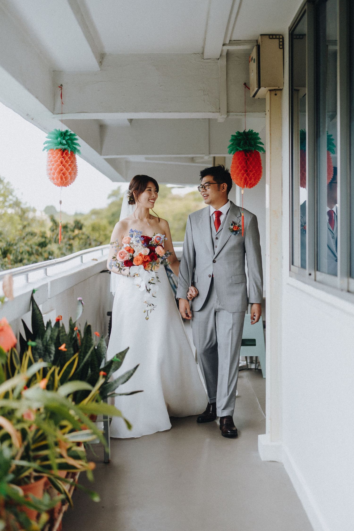 A couple in wedding attire walks down a tropical-themed balcony decorated with pineapple lanterns and plants.