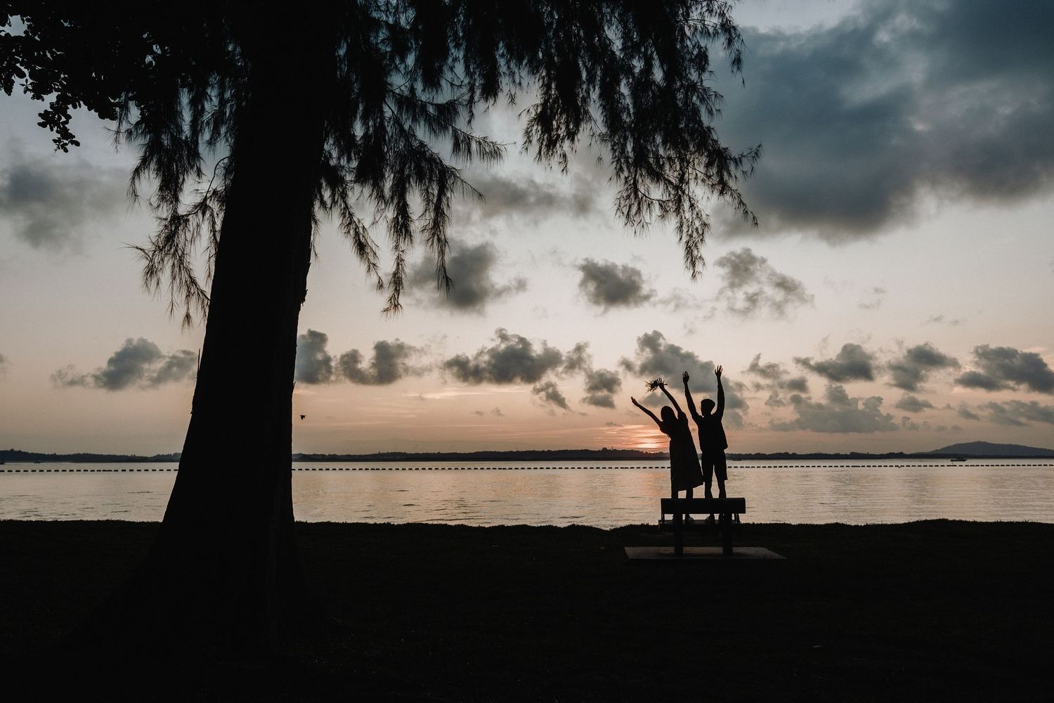 Two silhouetted figures sit on a bench under a tree at sunset with dramatic clouds in the sky.