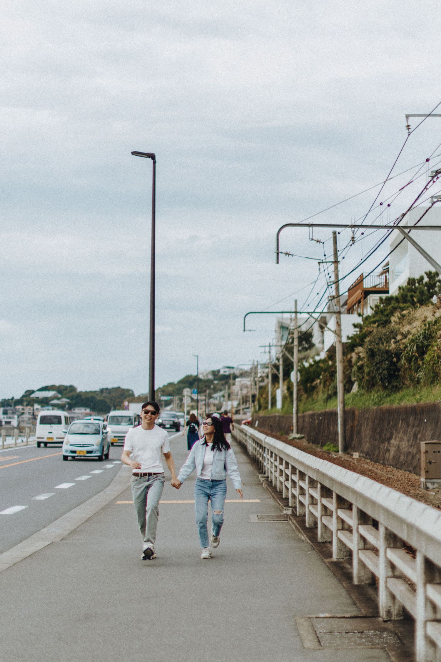 People walking along a coastal road with guardrails and utility poles on an overcast day.