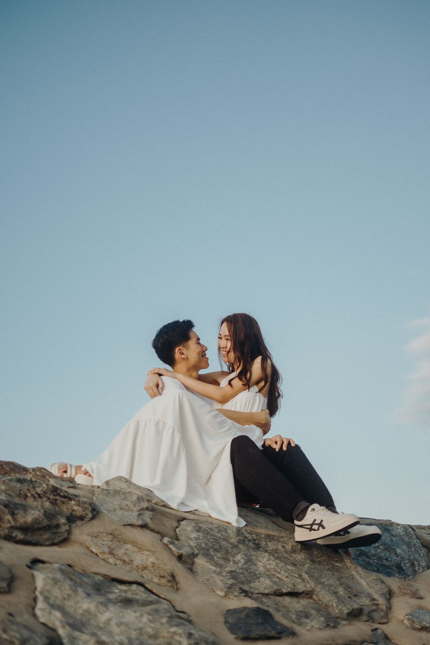 A couple embraces intimately while sitting on coastal rocks against a bright blue sky.