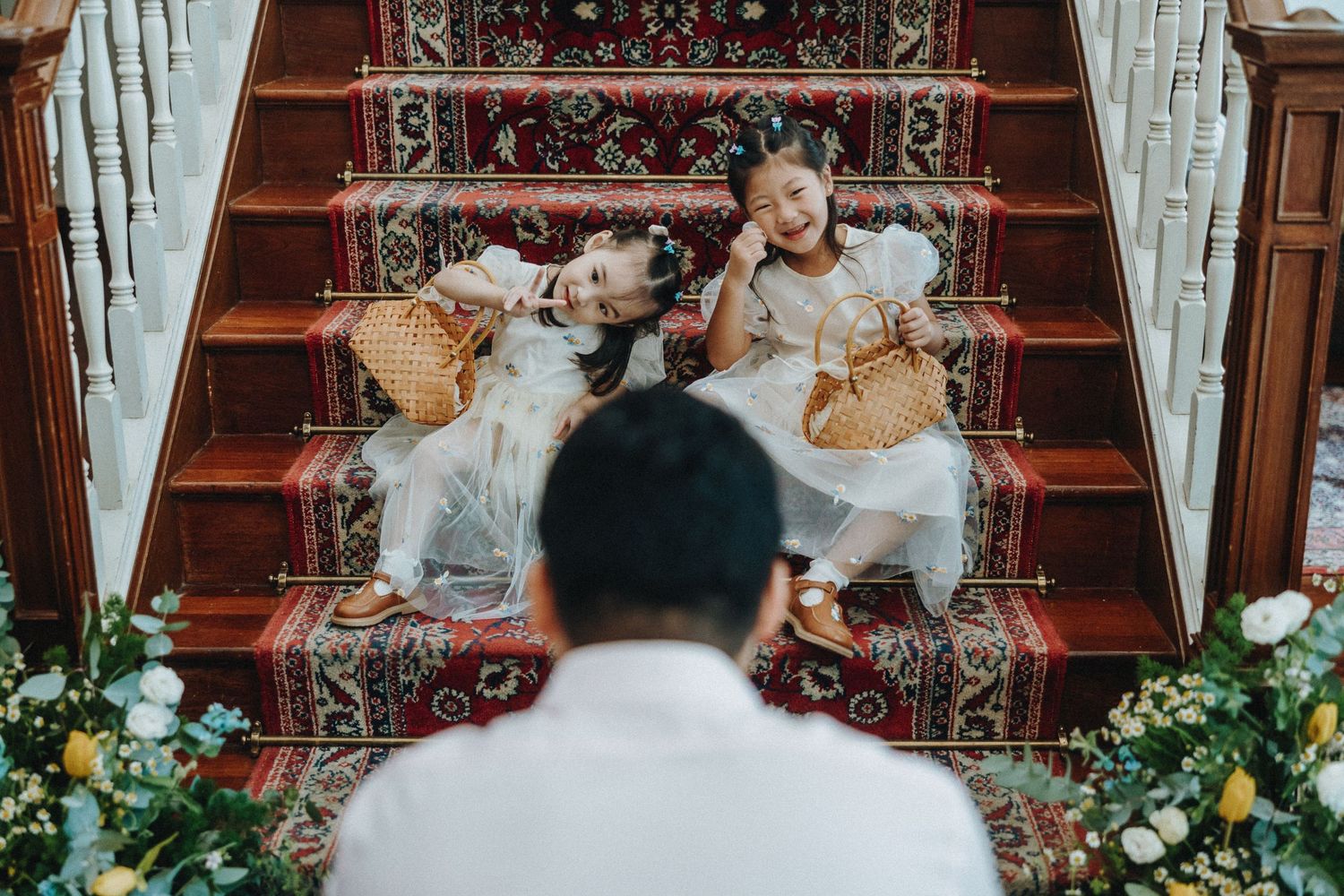 Two figures sit on elegant wooden stairs adorned with flowers while another looks up at them.