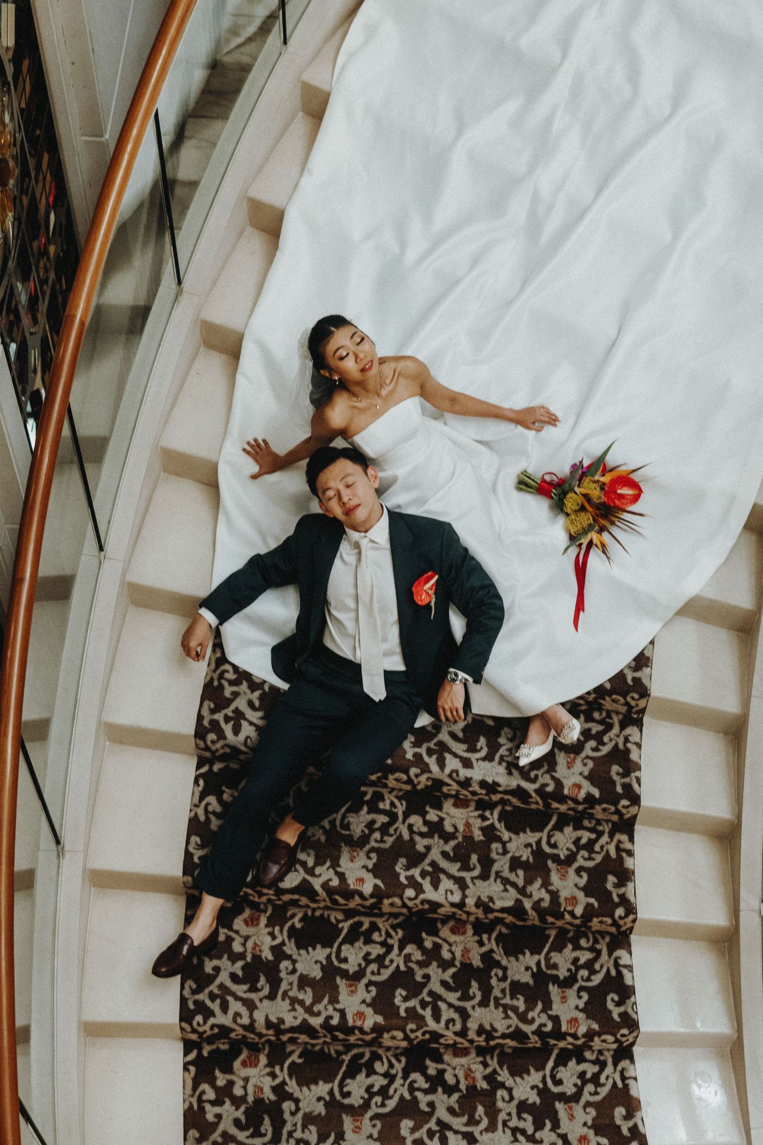 Creative overhead shot of couple laying on ornate carpeted staircase in wedding attire.