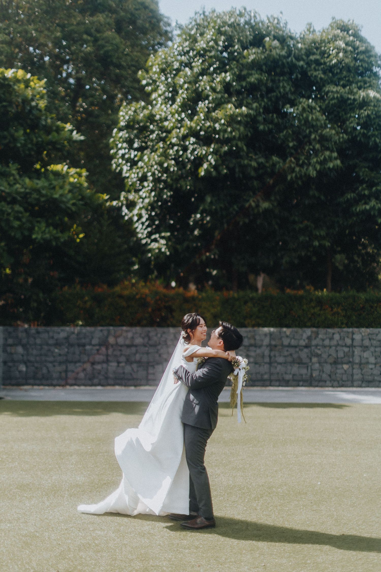 A romantic wedding couple dips and dances together on a lush green lawn with stone walls and trees in the background.