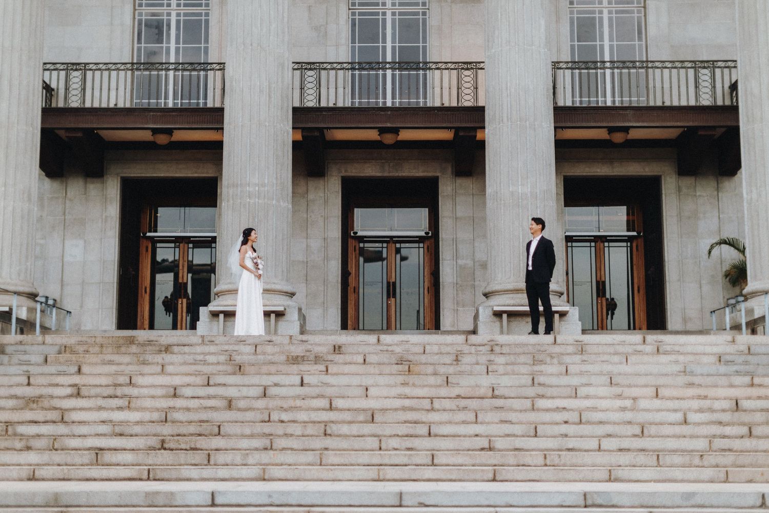 A couple in wedding attire poses on opposite ends of wide stone steps of a historic building.