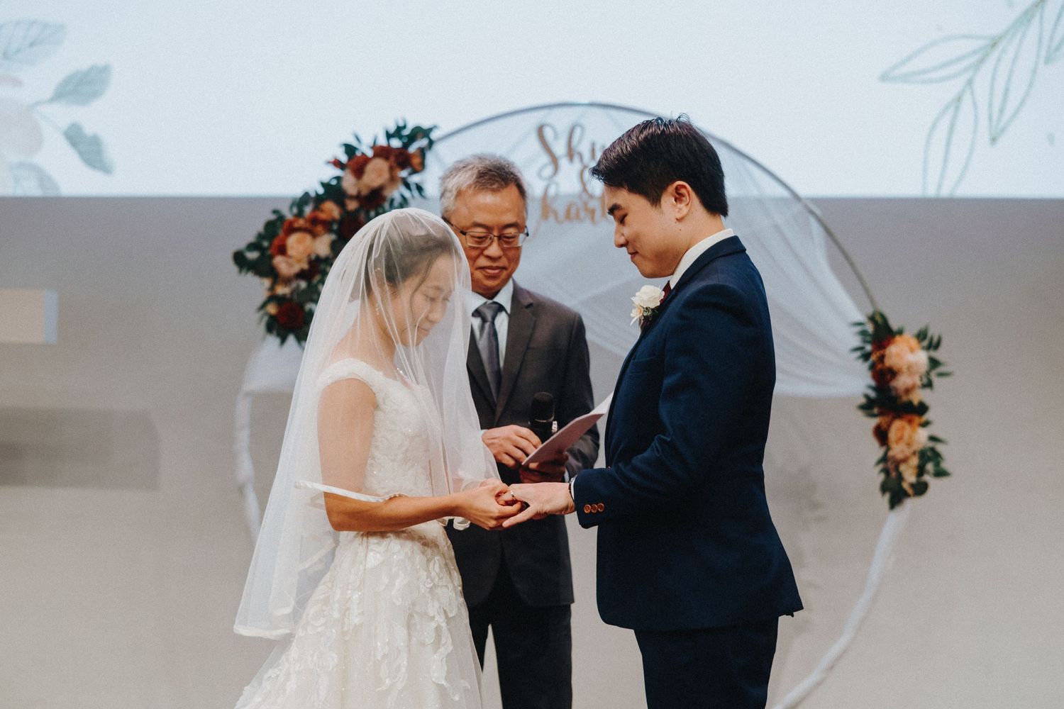 A wedding ceremony takes place under a circular arch decorated with peach and coral flowers.