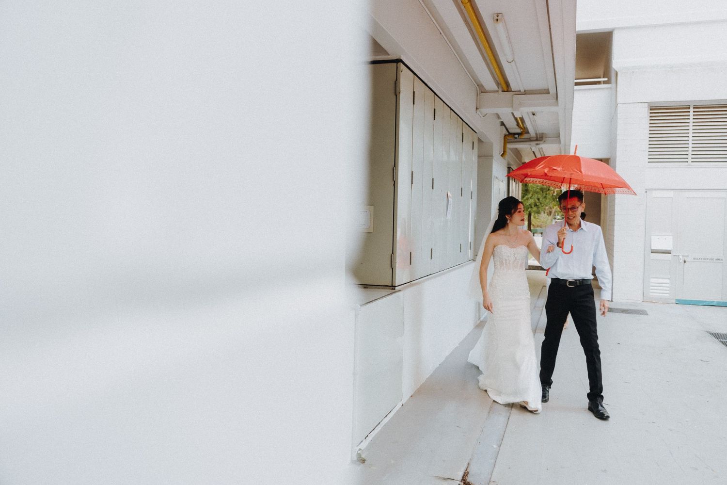 Two figures stand in a bright white hallway with one holding a bright red umbrella.
