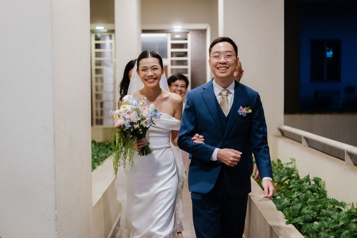Smiling Asian bride and groom walk arm in arm down an outdoor corridor, bride holding a lush floral bouquet.