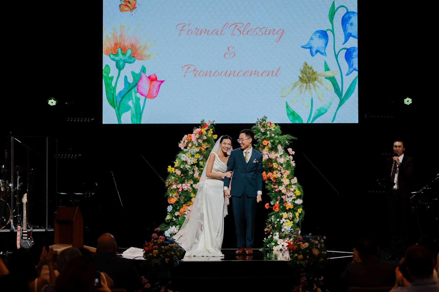 Bride and groom standing under floral arch at formal blessing and pronouncement wedding ceremony on stage.