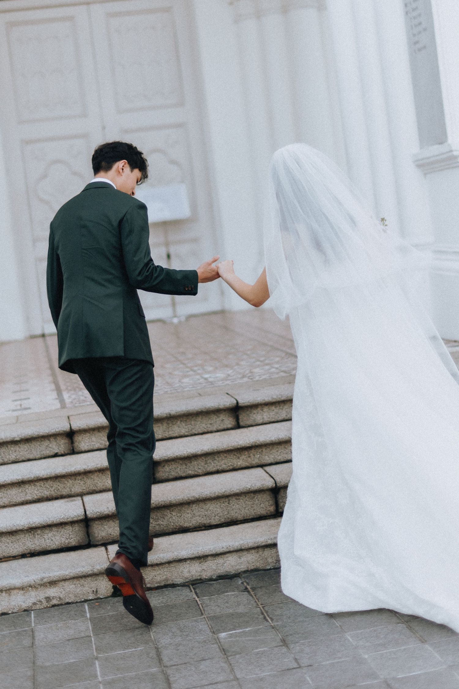 Bride and groom holding hands walking up church steps, groom in dark suit, bride in white gown with veil.