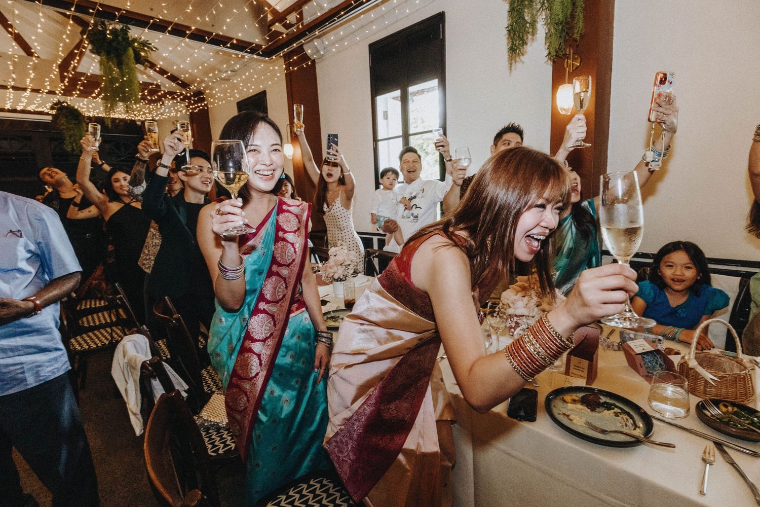 Guests in colorful saris celebrating at an indoor wedding reception, raising glasses of wine and smiling joyfully.