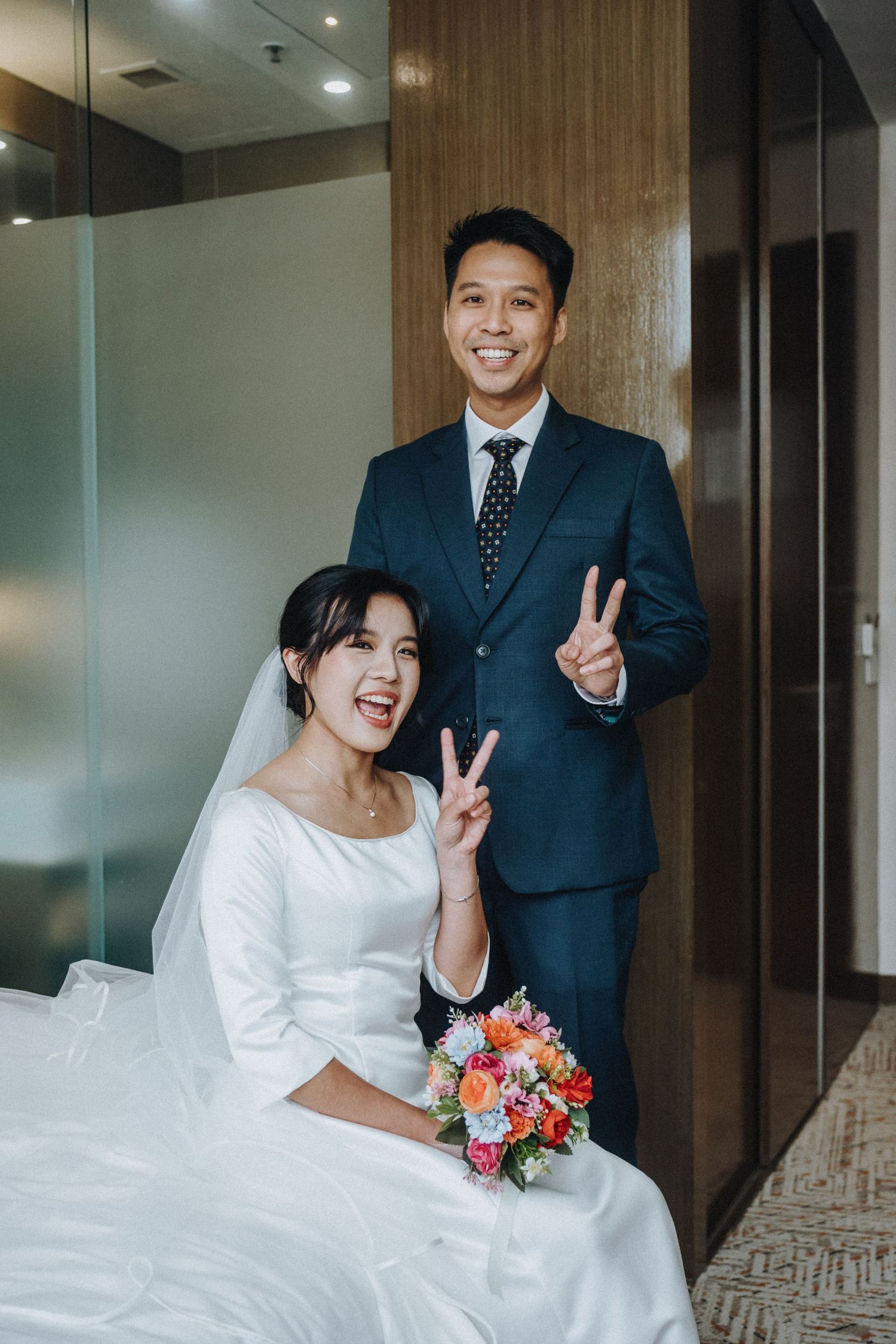 Happy Asian bride and groom in wedding attire making peace signs, bride holding colorful bouquet.
