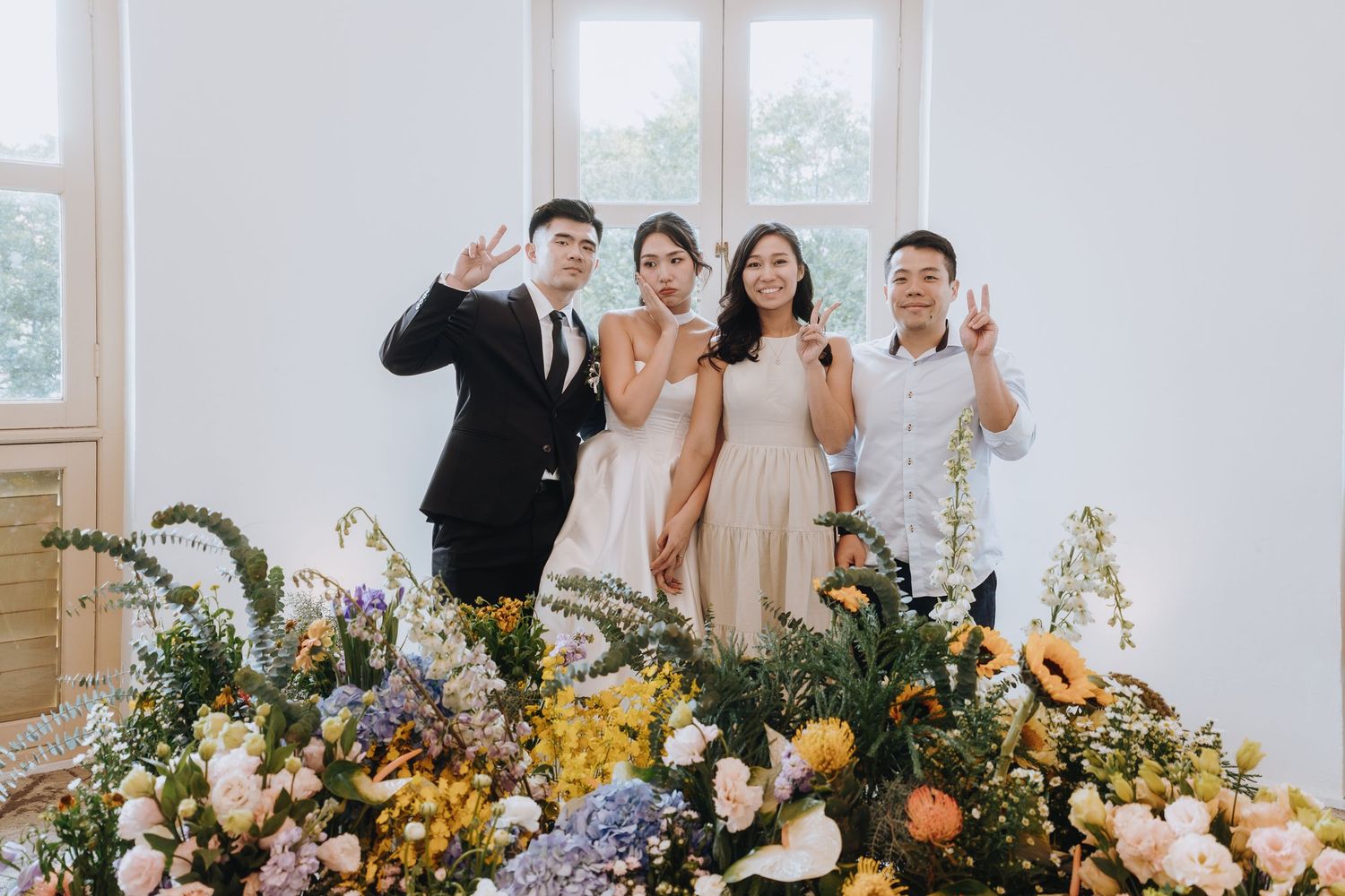 Four guests posing with peace signs beside colorful floral arrangements at a wedding.