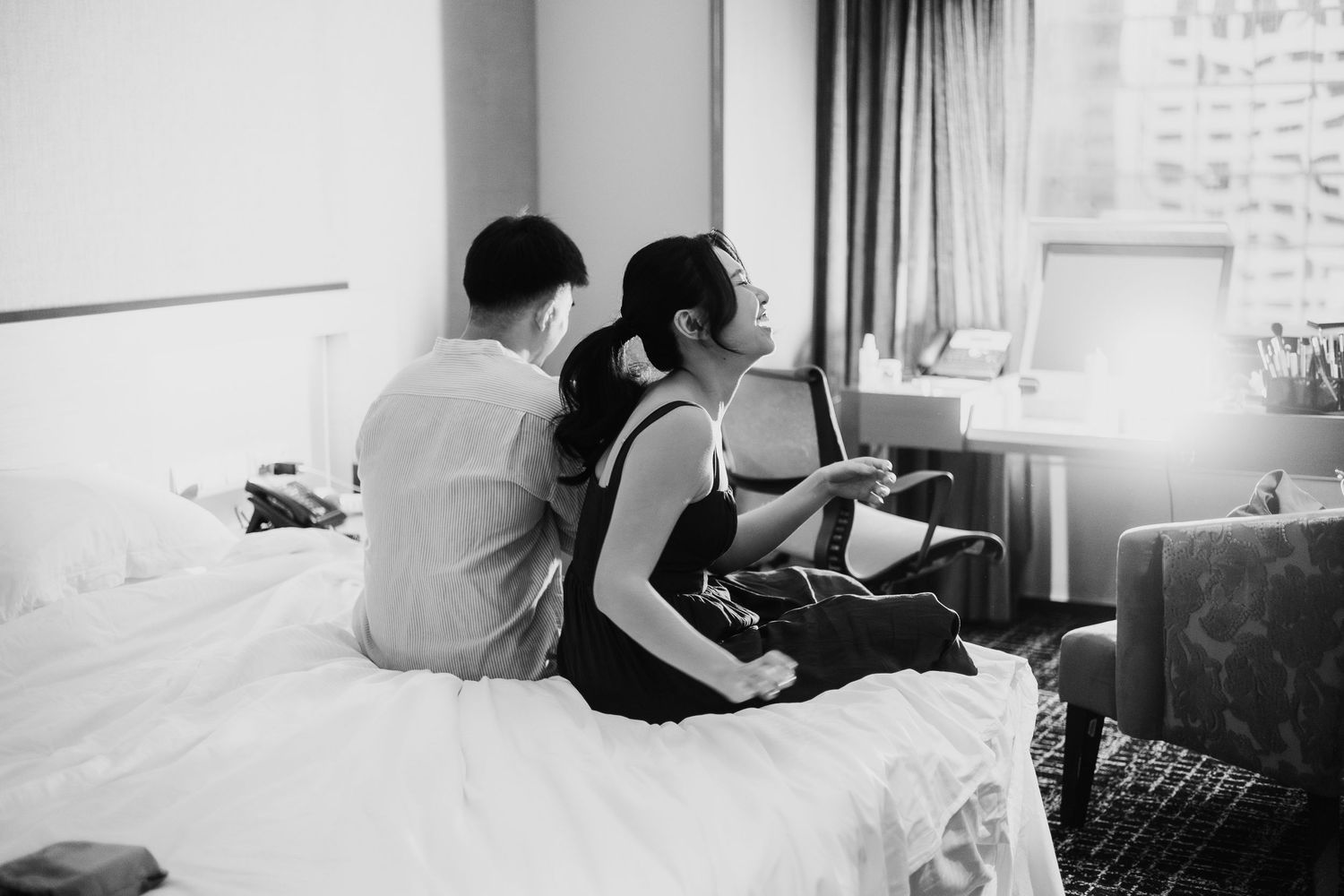 Couple sitting together on a white bed in a hotel room, captured in black and white photography.