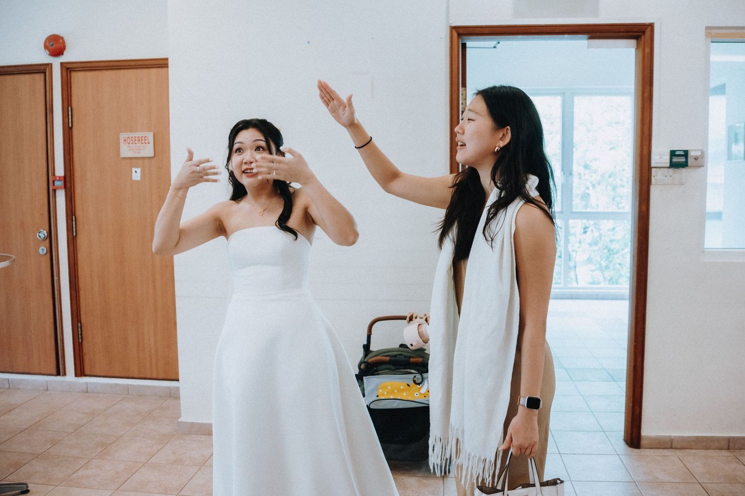 Bride in white strapless gown gestures with woman in white dress during wedding preparation indoors.