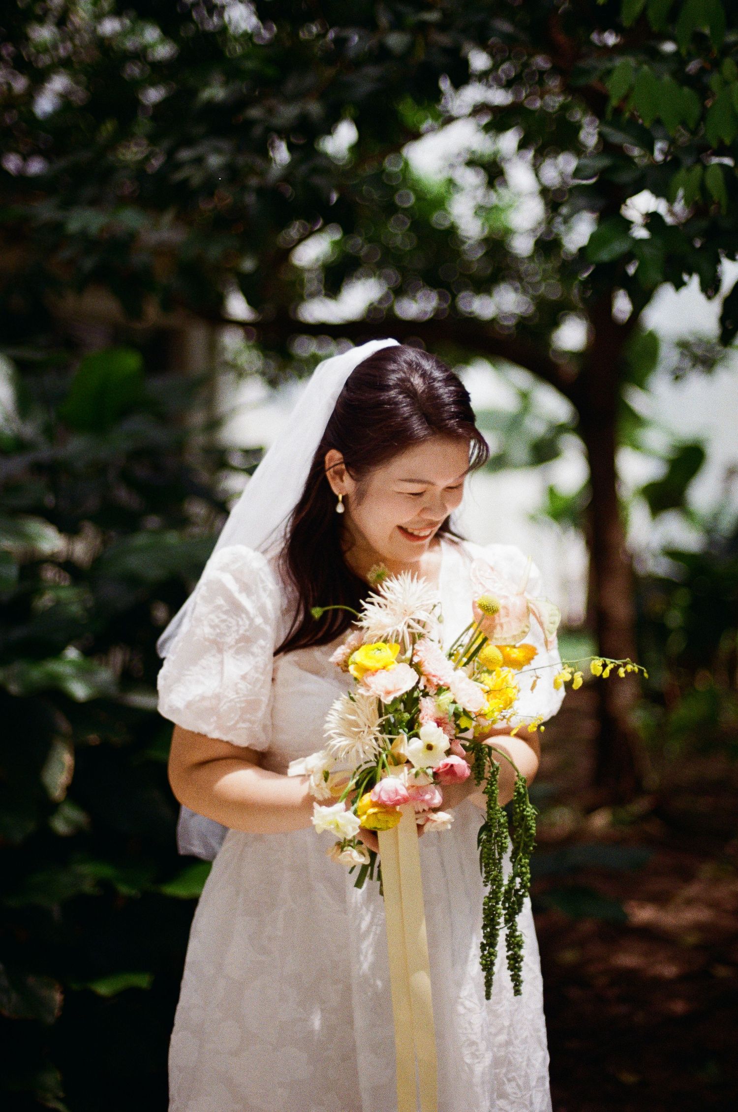 Woman in white lace dress holding a bright yellow and pink floral bouquet in a sunlit woodland setting.