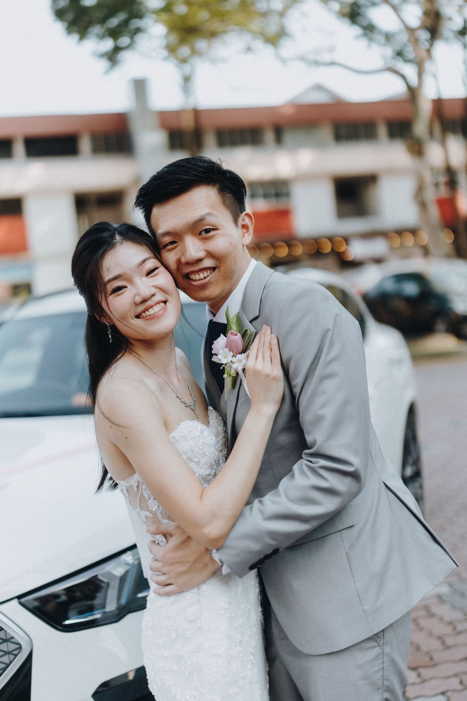 A joyful wedding couple embraces next to a white car in their elegant wedding attire.