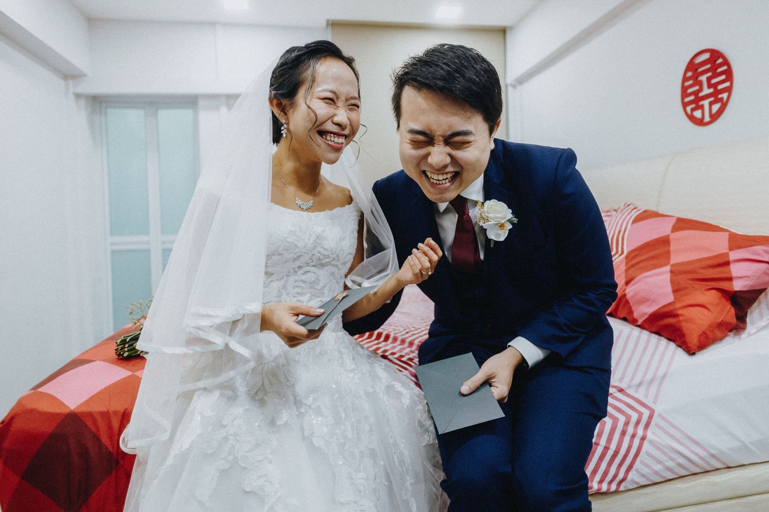 A joyful wedding couple share a laugh while sitting on red and white cushions at home.