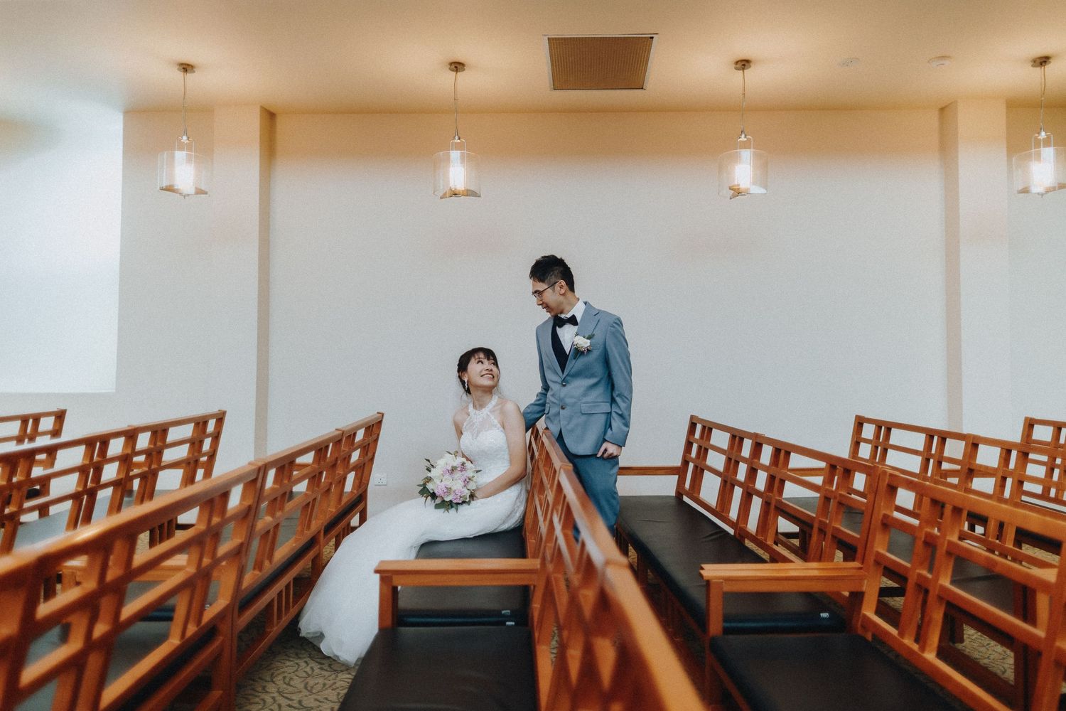 A couple in wedding attire shares an intimate moment among wooden church pews in a minimalist chapel.
