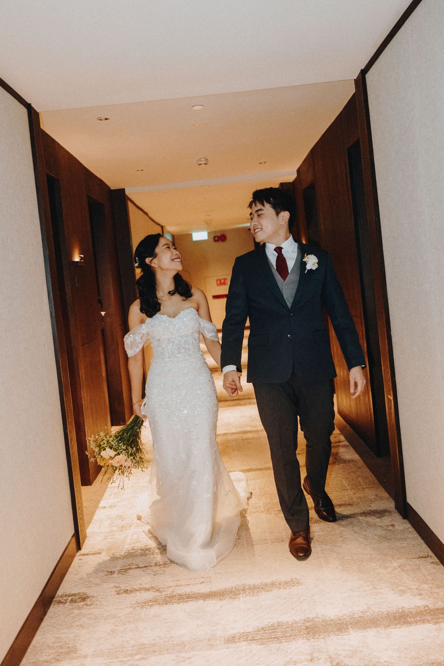Newlyweds walking down a hallway, bride in white dress and groom in dark suit.