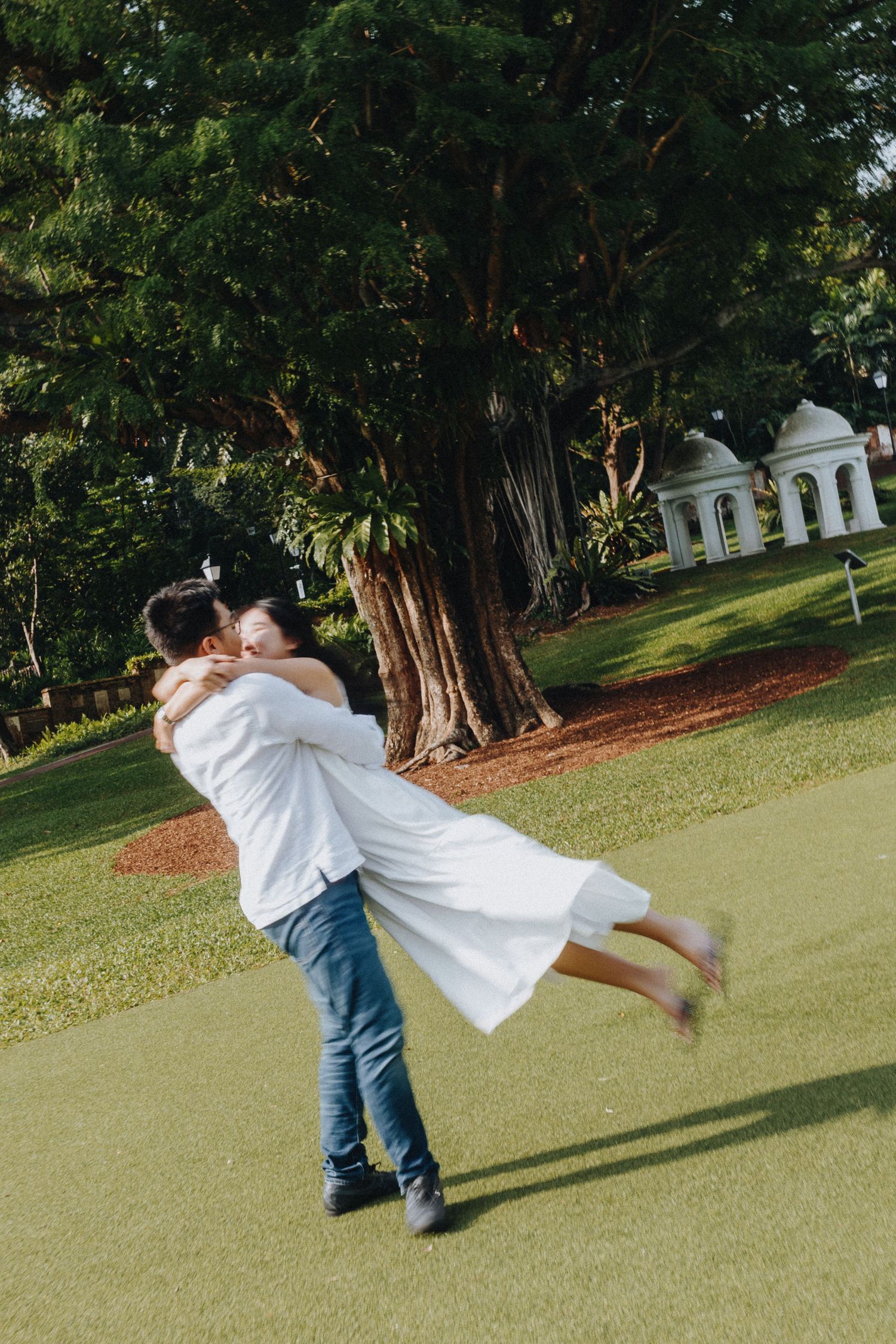 A joyful couple embraces in a spinning motion on a green lawn with a large tree and gazebo behind them.