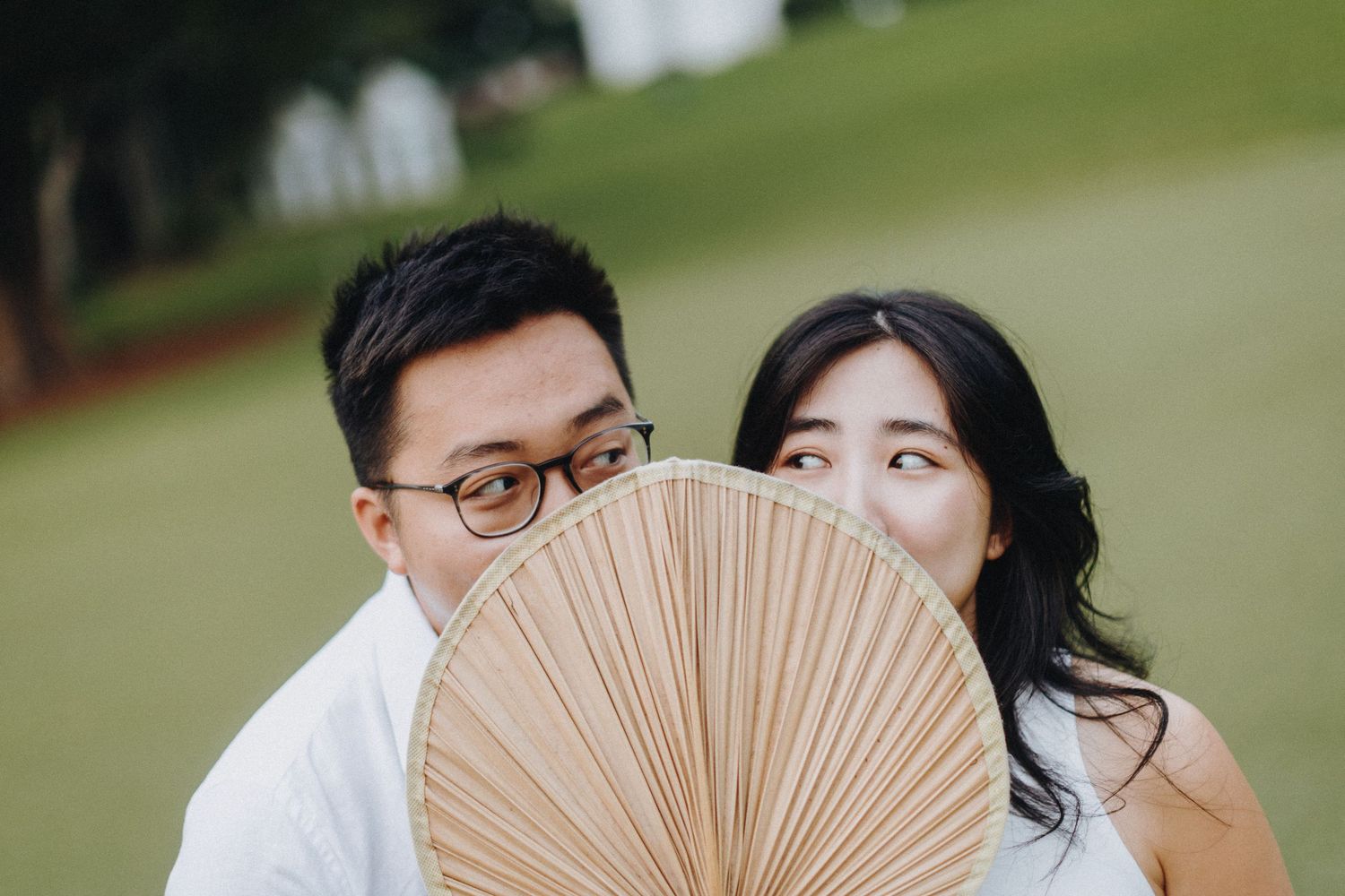 A couple shares a playful moment behind a tan paper fan in an outdoor garden setting.