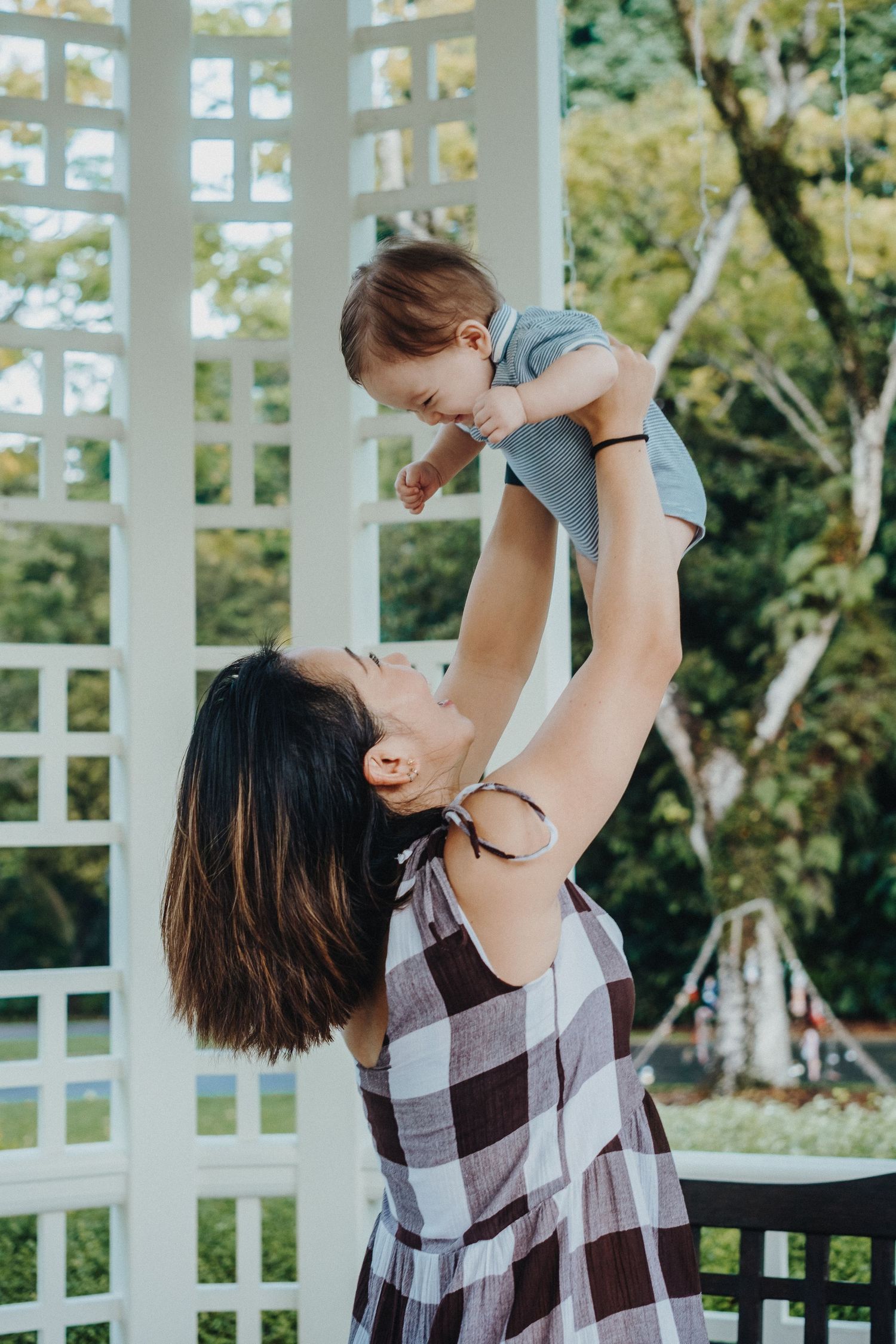 A mother lifts and plays with a young child in a white gazebo surrounded by lush greenery.