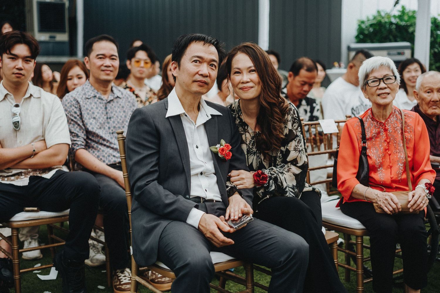 Wedding guests seated outdoors smile while watching ceremony from wooden chairs at outdoor venue.