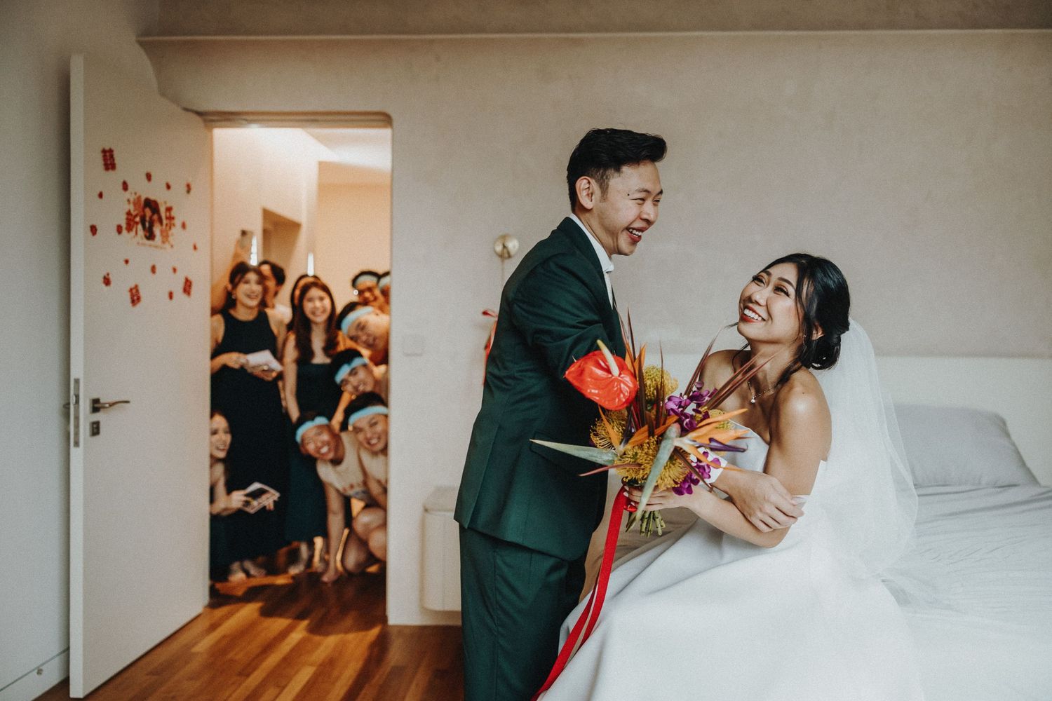 Wedding couple shares a joyful moment while guests peek through doorway during intimate indoor celebration.