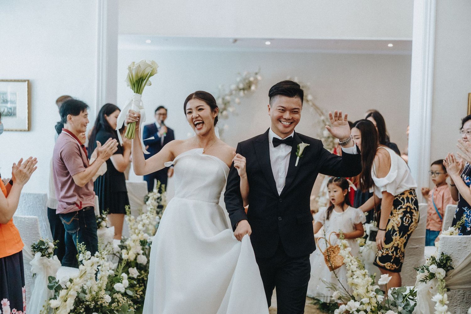 Newlyweds celebrate while walking down an aisle decorated with white flowers as guests applaud.