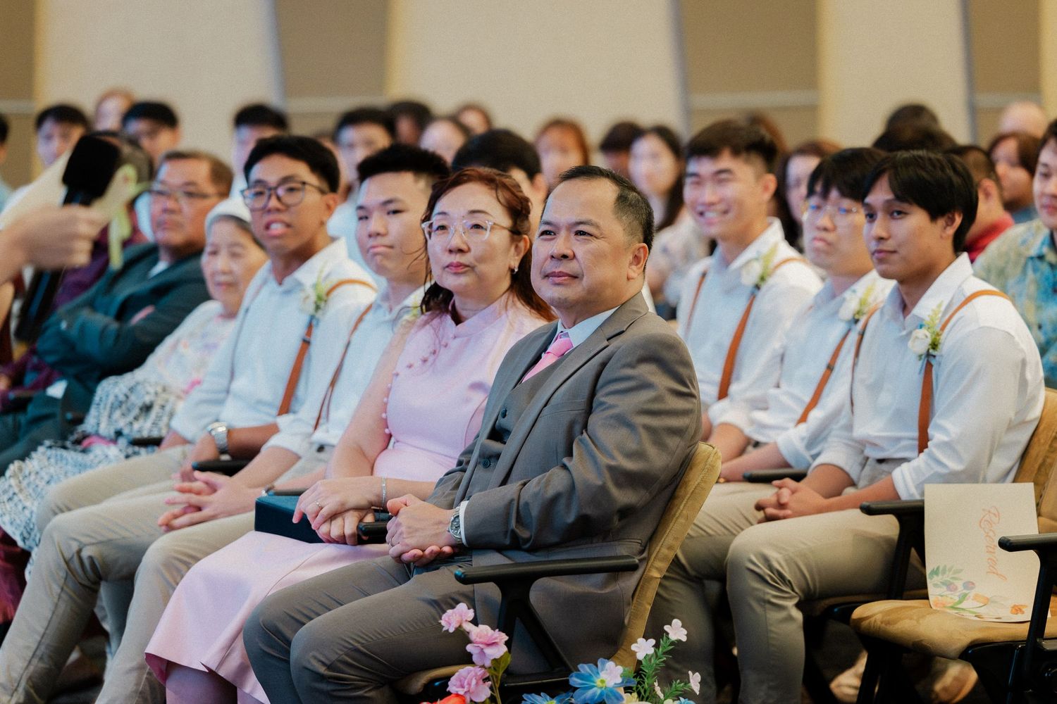 Smiling wedding guests including parents seated in rows at elegant wedding ceremony, wearing formal attire with floral corsages.