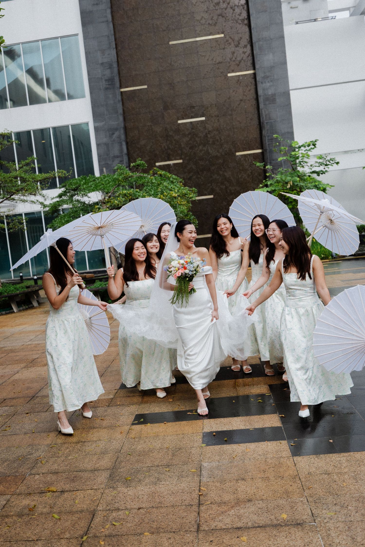 Bride and bridesmaids in white dresses holding parasols laughing outdoors on rainy wedding day.