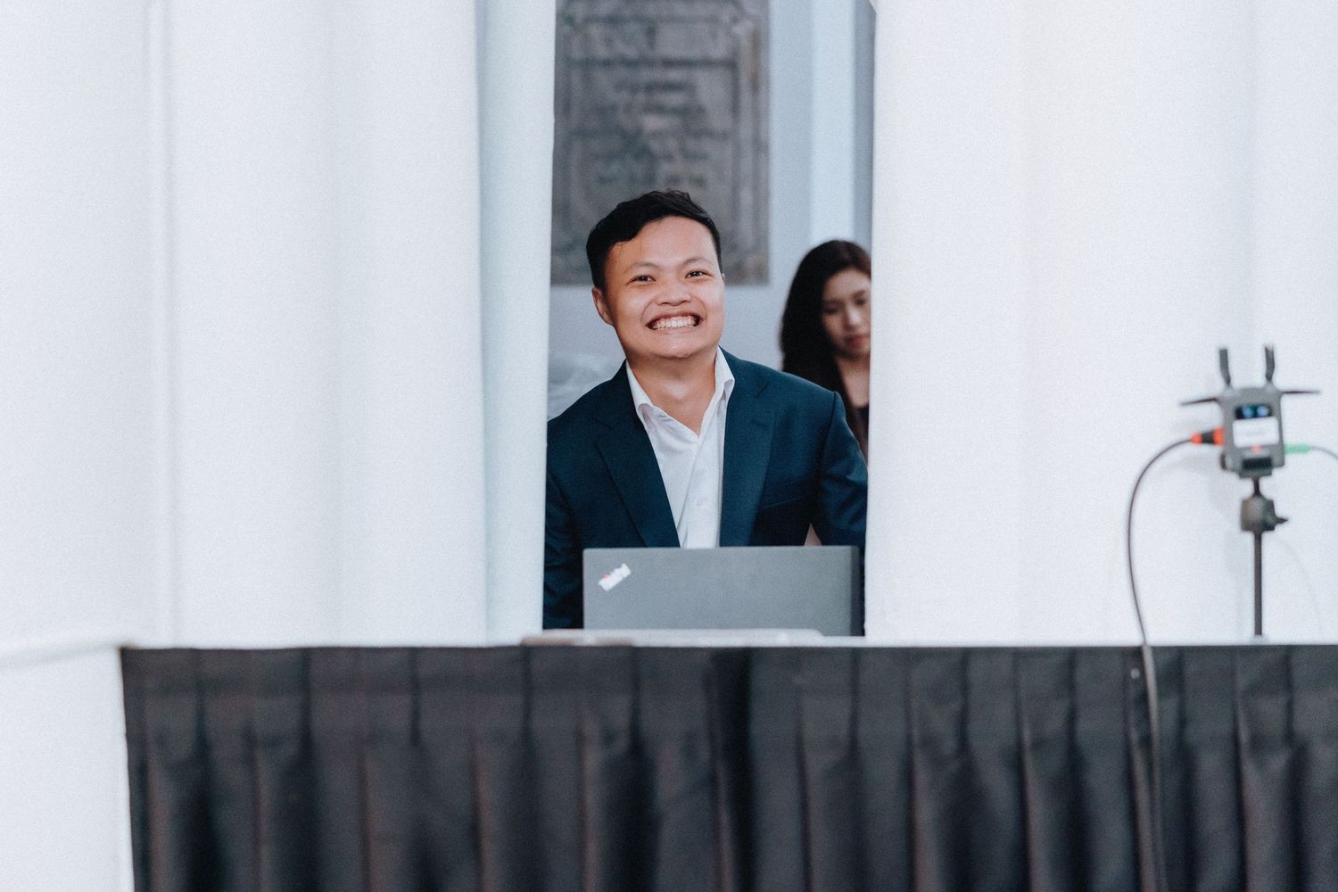 Smiling Asian man in a dark blazer standing behind a laptop at a formal event venue.