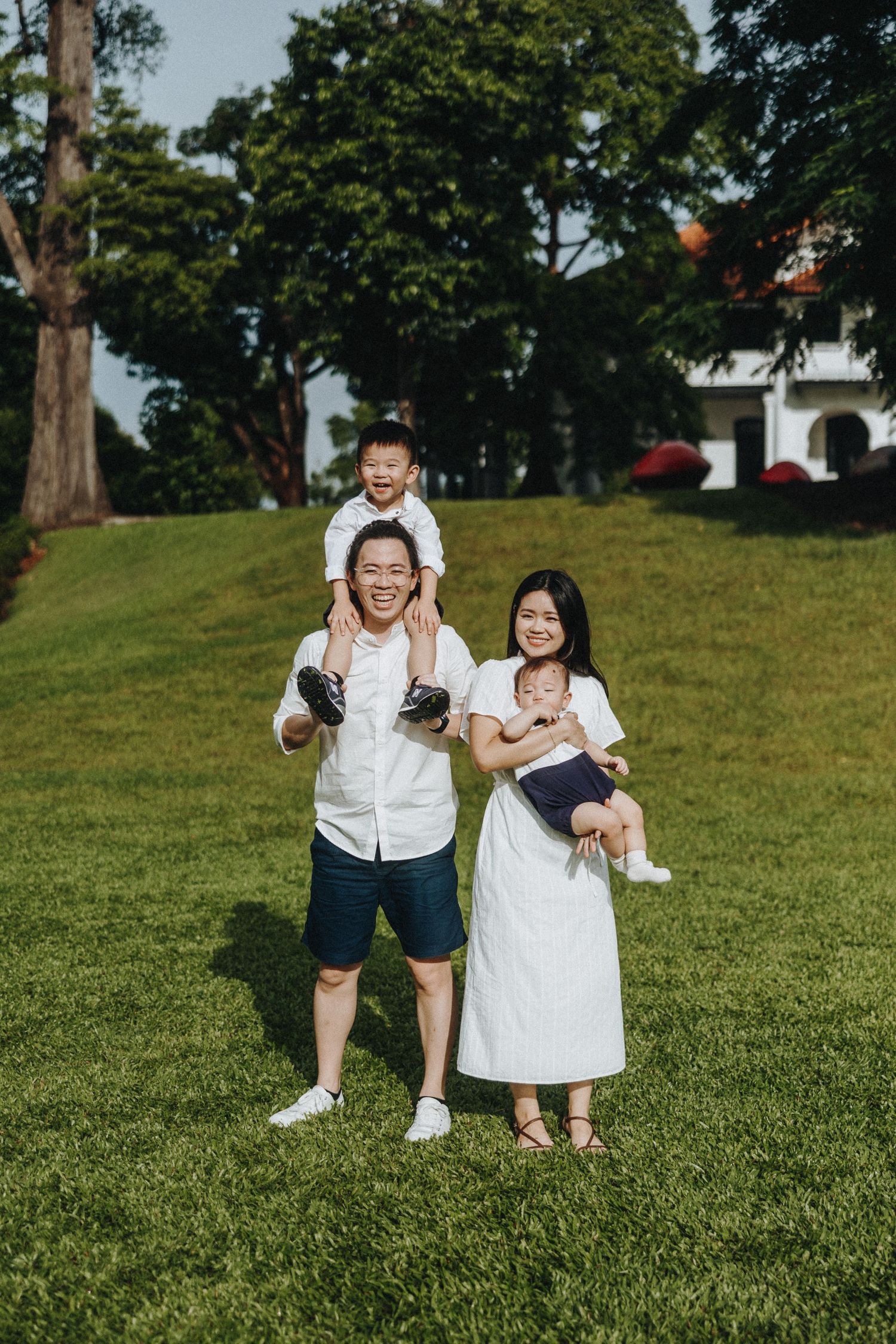 Family wearing white outfits poses together on a grassy hill with trees and a white building in the background.