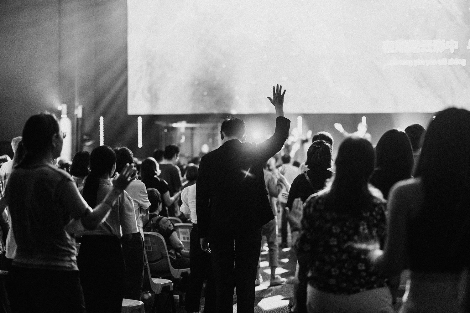 Black and white shot of silhouetted audience members with raised hands at a worship service.