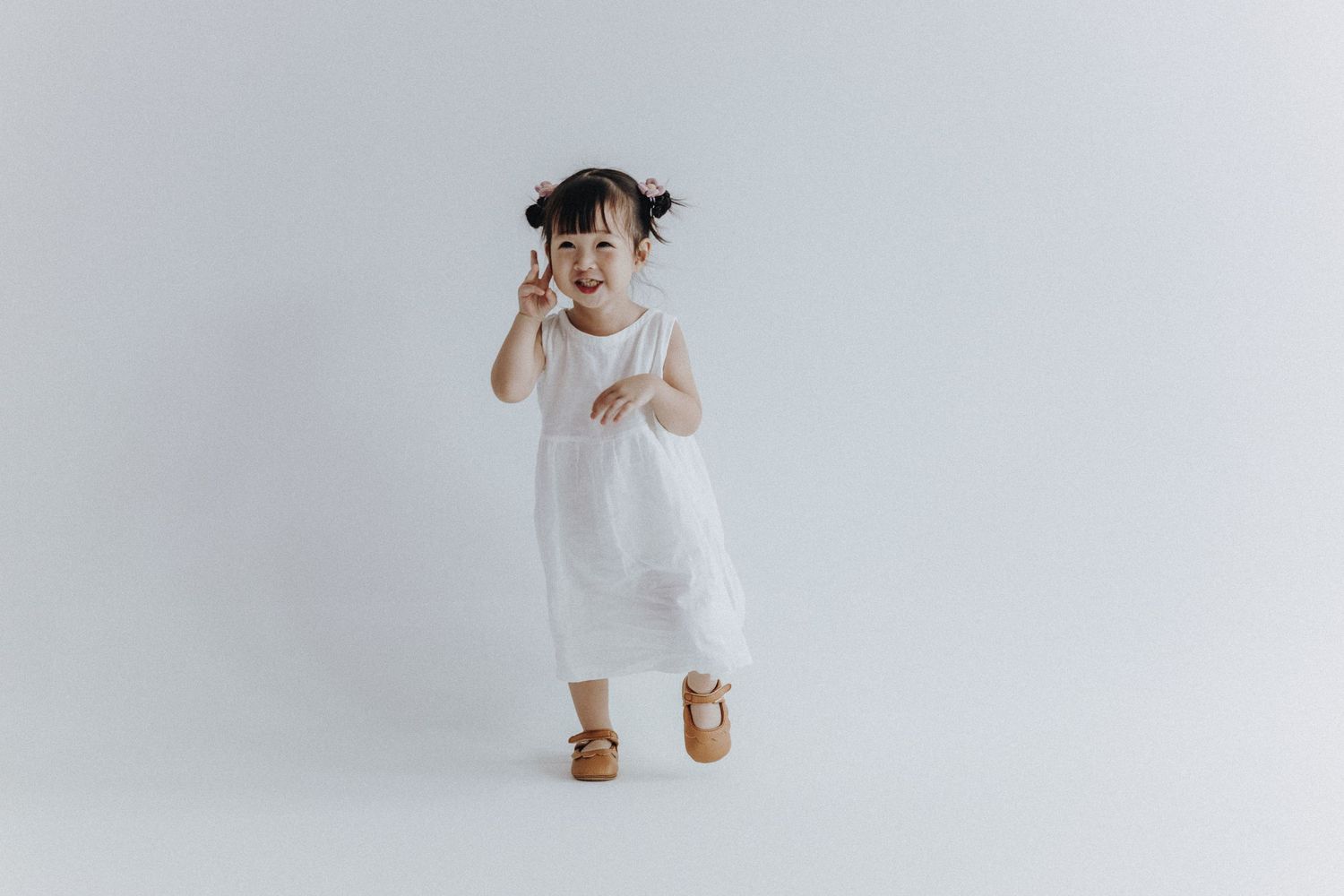 Young child in white dress making expressive gesture against minimal white background.