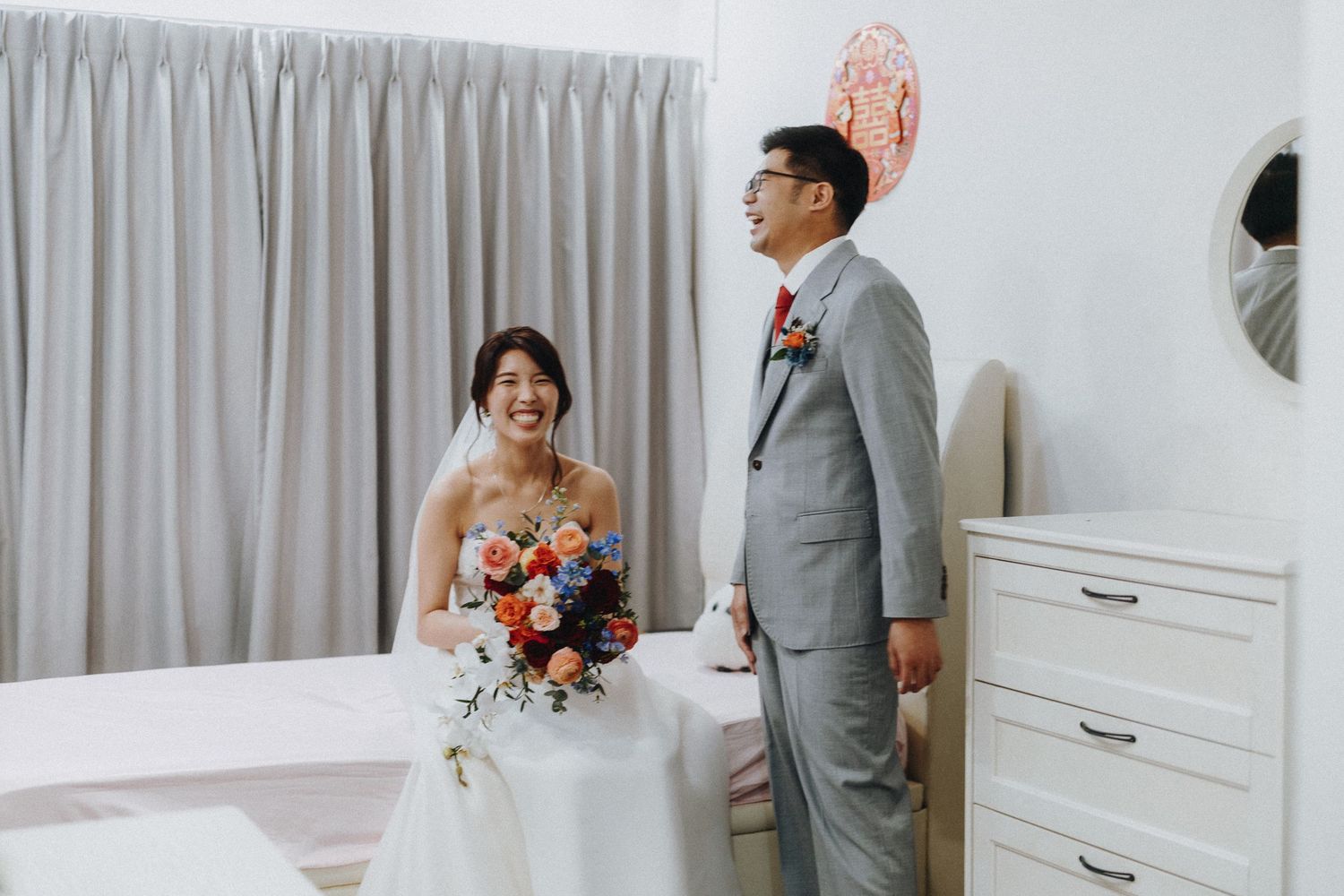 Bride and groom pose together in a bright room, with the bride holding a vibrant orange and pink bouquet.