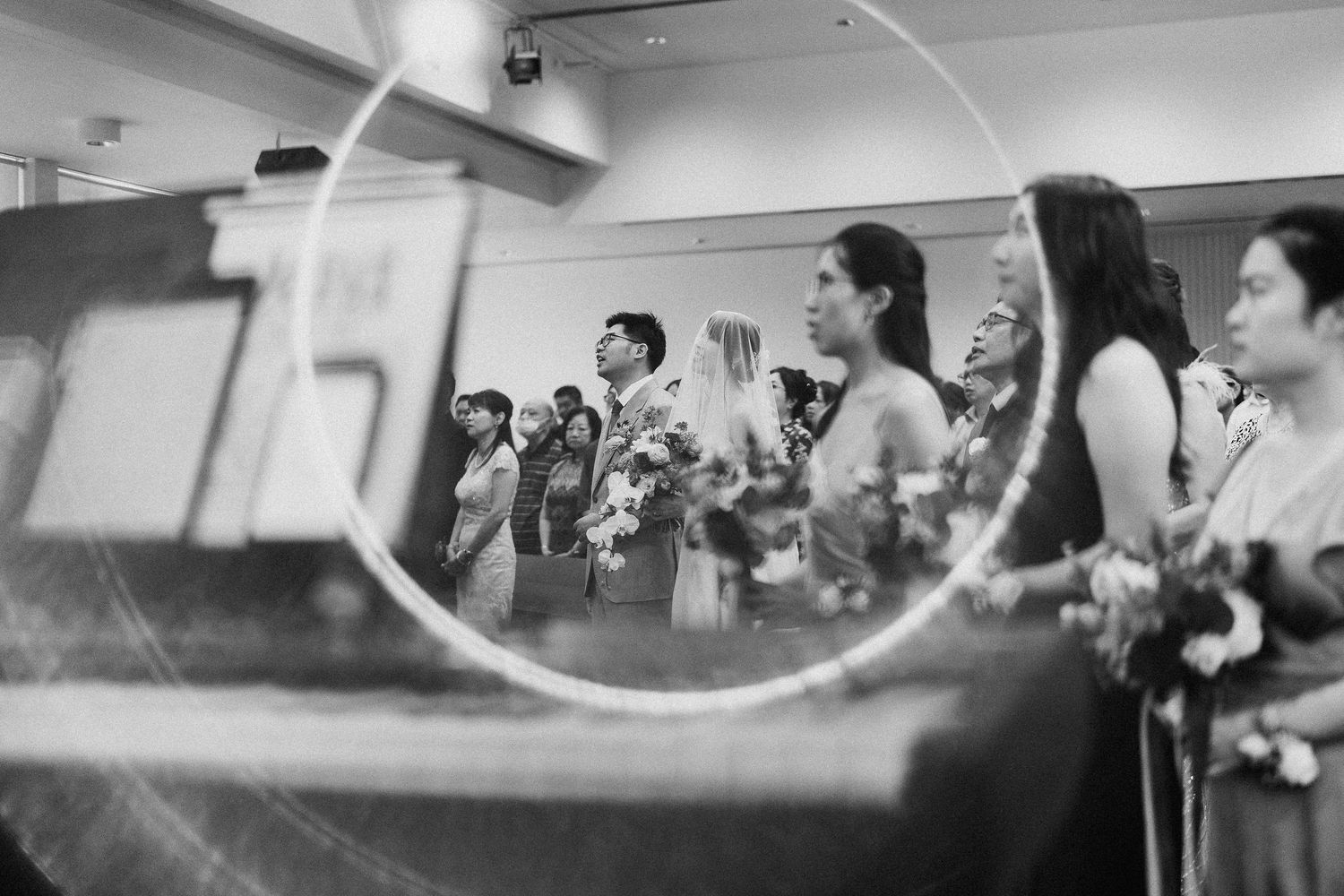 A black and white reflection through glass shows wedding attendees holding bouquets.