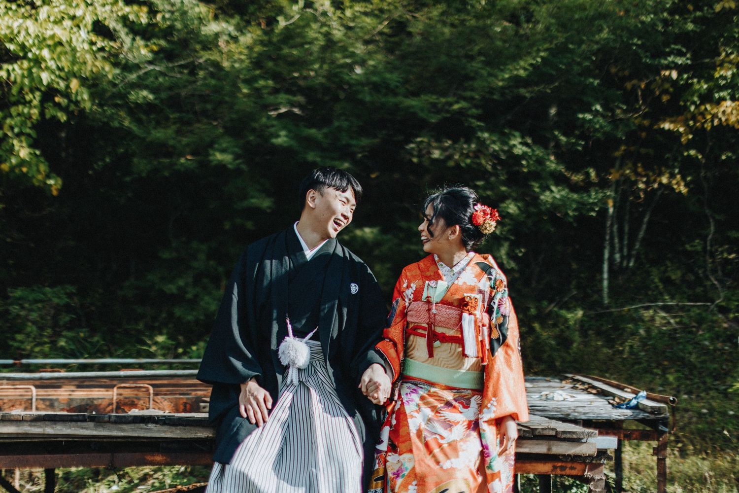 Couple in traditional Japanese attire poses together in a lush garden setting with autumn foliage.