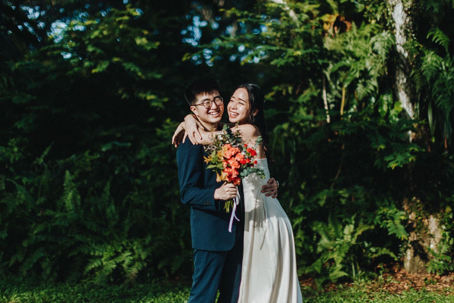 A couple embraces during an outdoor wedding photoshoot surrounded by lush green foliage.