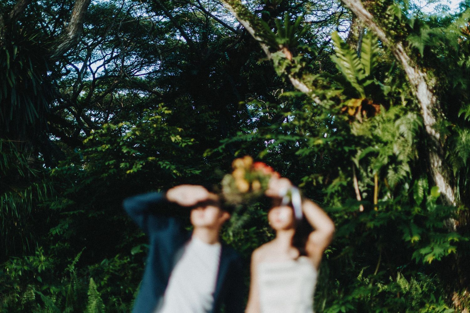 Wedding couple poses together in lush garden setting with dappled sunlight filtering through trees.