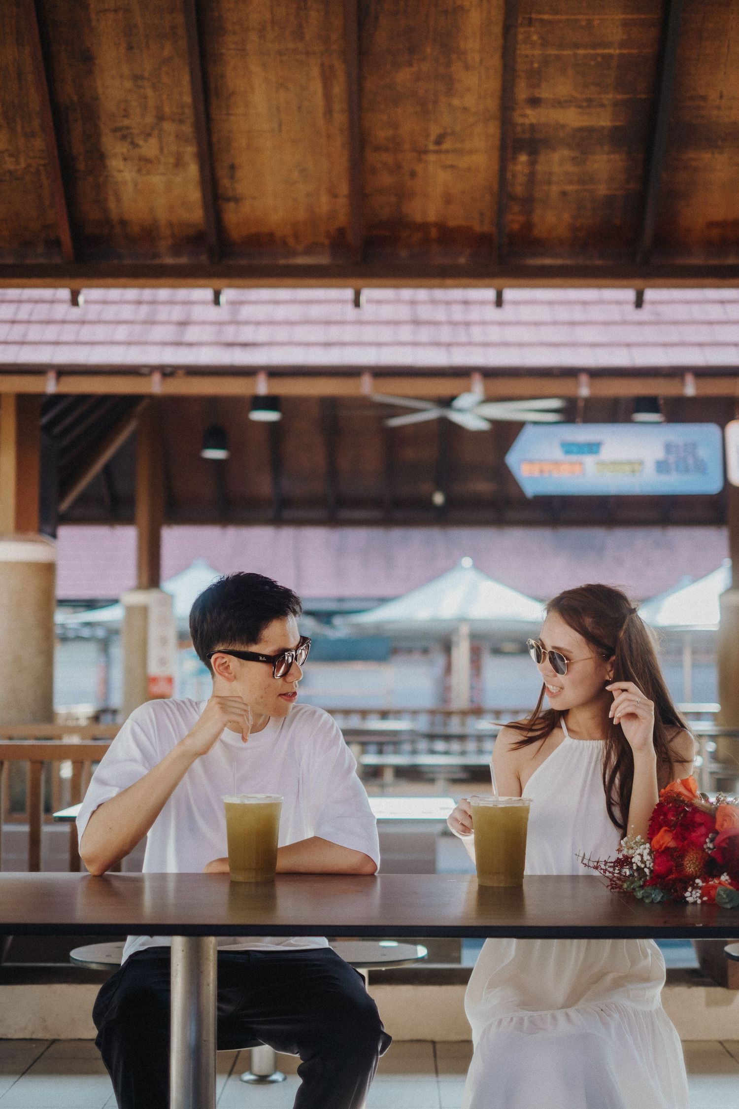 Two people sitting at outdoor cafe tables with drinks and red flowers.