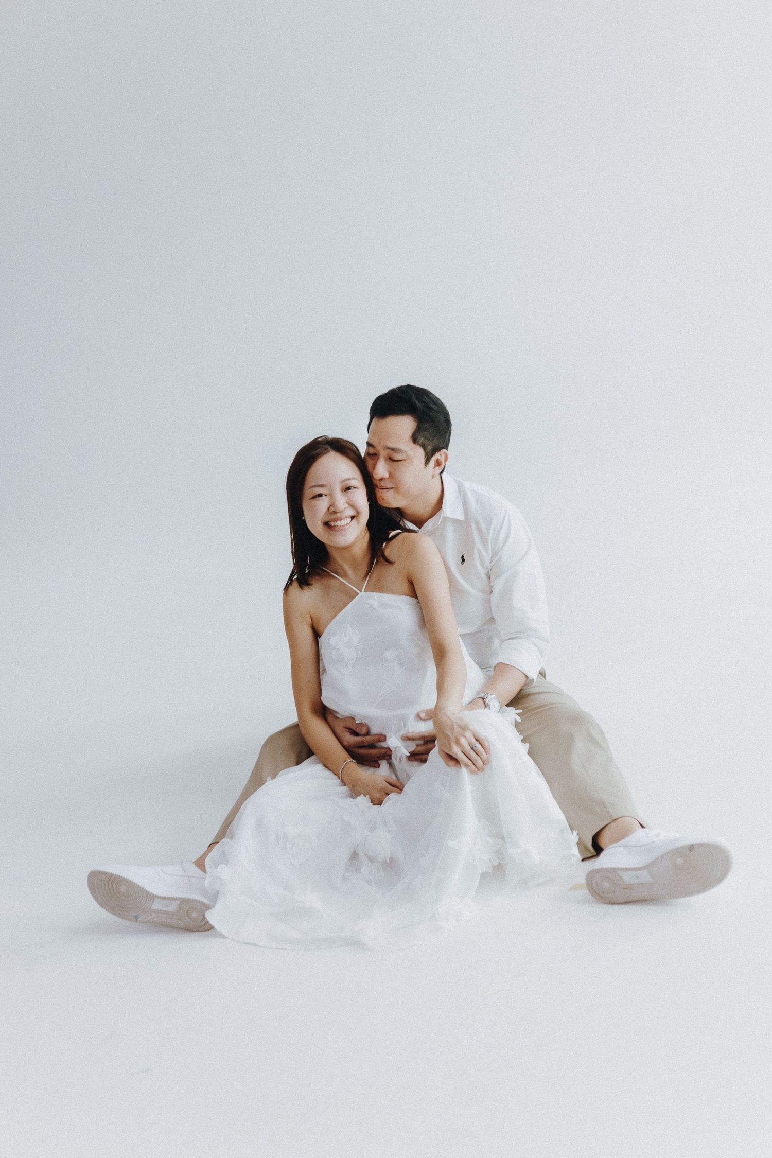 A couple in white attire sits together intimately against a plain light background during a romantic photoshoot.