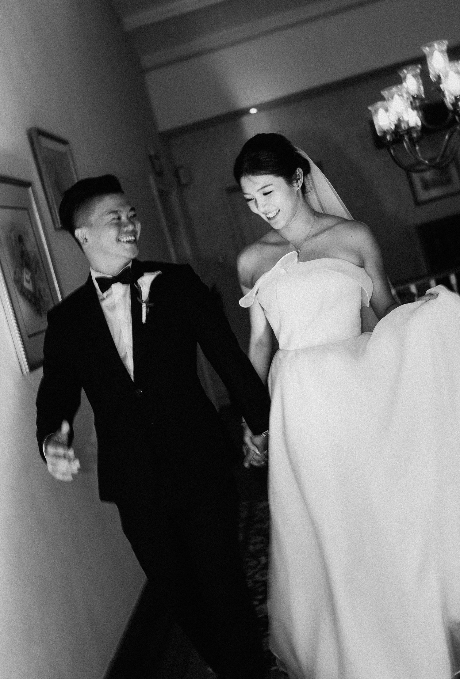 A bride in an elegant off-shoulder gown and groom in tuxedo share a joyful moment walking together in black and white.