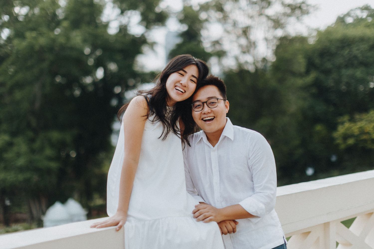 A joyful couple in white outfits share a candid and loving moment while sitting together on a railing outdoors.
