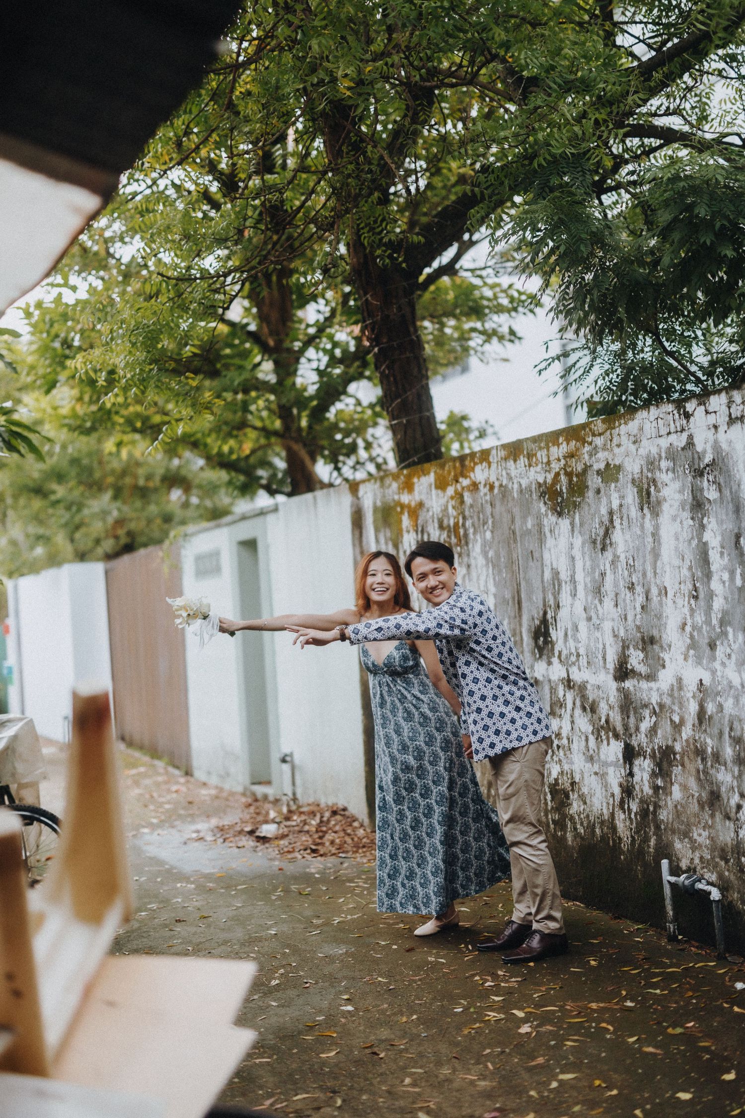 Duo posing and dancing by weathered white wall in garden setting.