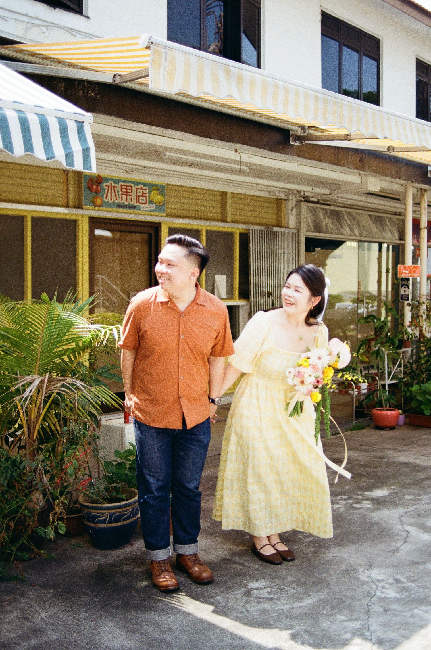 A couple shares a joyful moment outside a shop with yellow awnings and tropical plants.