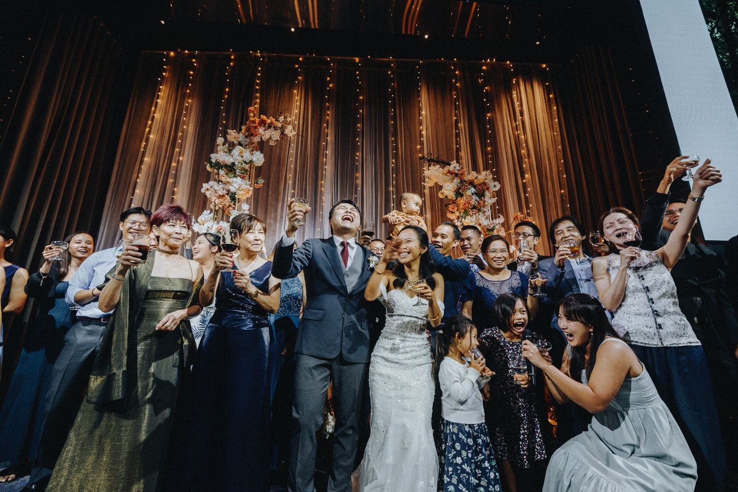 Joyful wedding celebration shot with guests cheering and raising their hands against a lit backdrop.