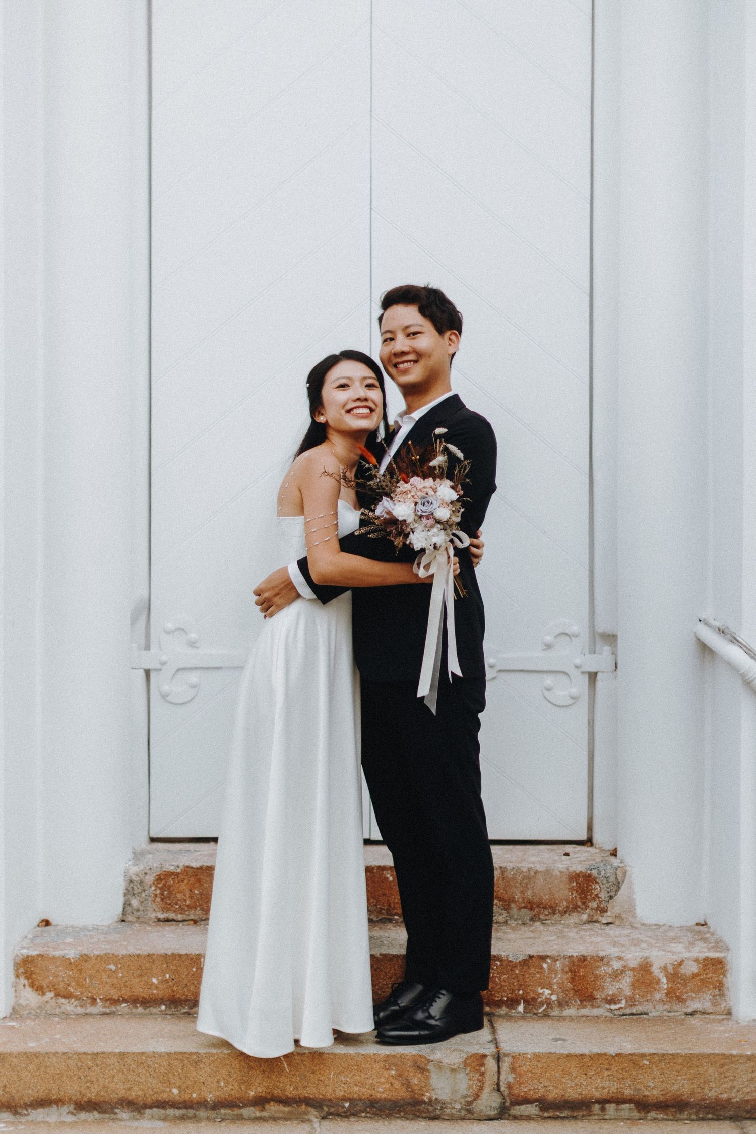 Elegant couple in formal attire posing on white steps with bouquet.